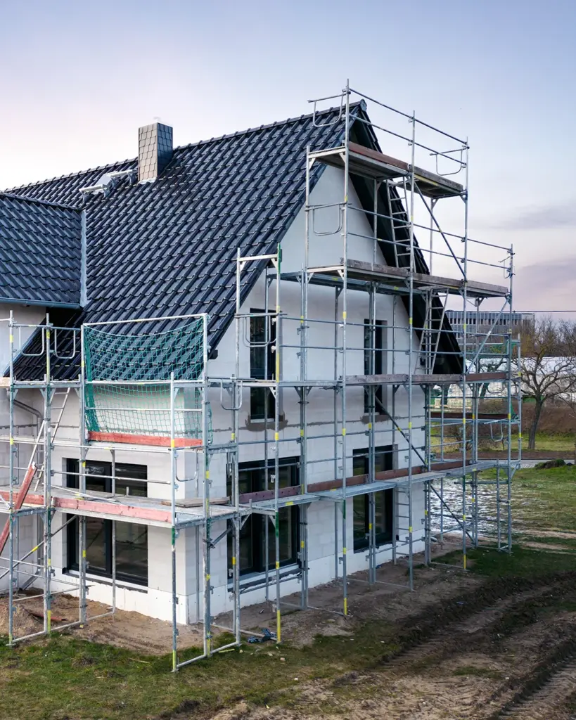 Sinnbild für Unterstützung von Baugutachten durch Heike Schwartz. Das Bild zeigt einen Neubau mit einem Baugerüst an der Fassade. Das Dach ist mit blauen Dachziegel gedeckt und die Fassade weiß. Die Rahmen der Fenster sind schwarz.